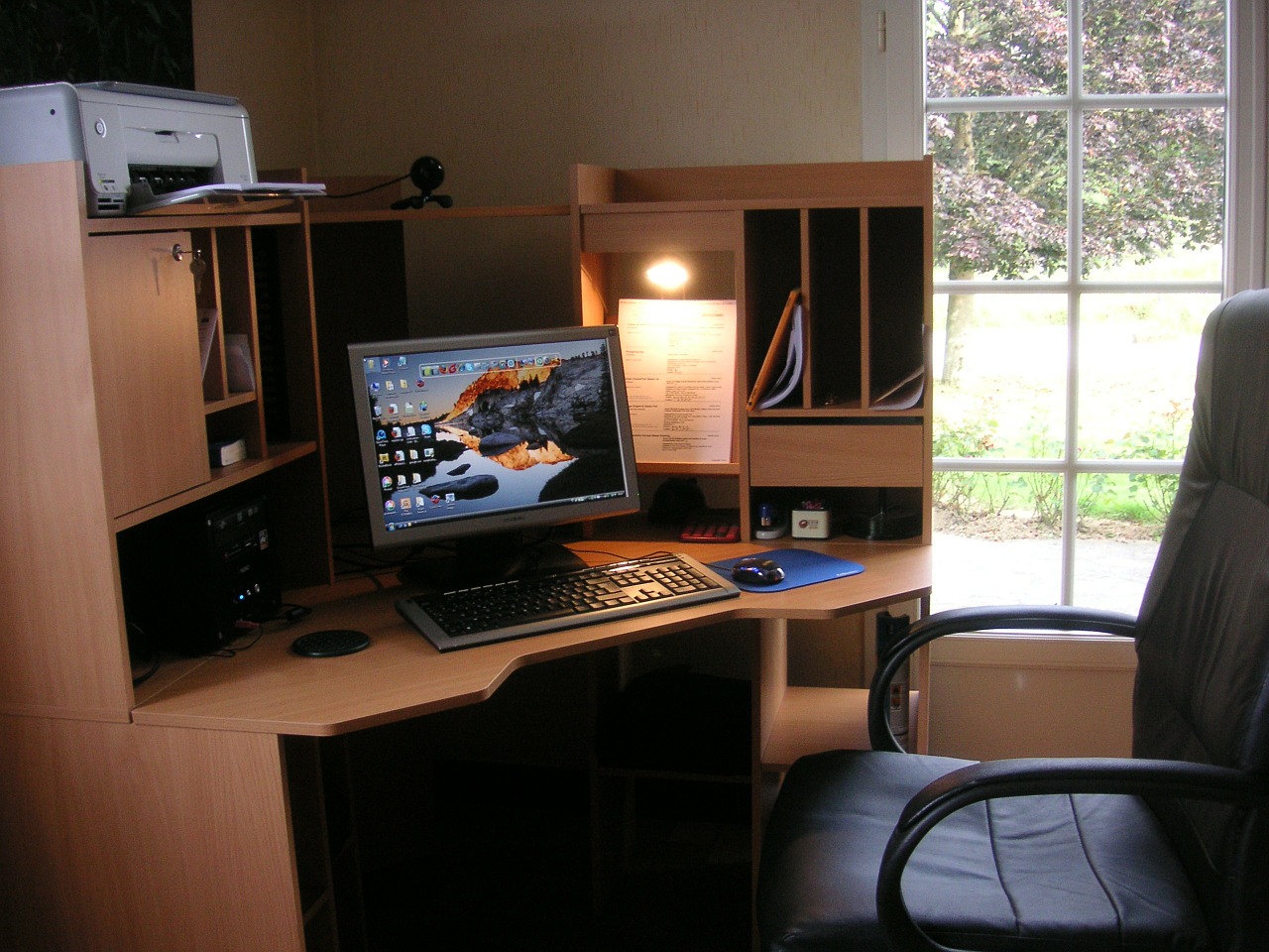 An office desk with computer monitor keyboard and office chair sitting empty.  Large window to the right appears to be a home-office set-up. 